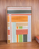 Quadro decorativo Enlace Prisma de 30x20 cm em madeira e linho, com fios em blocos de rosa, verde oliva, laranja, terracota e lilás, em moldura de madeira, apoiado em estante de madeira.