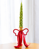 Castiçal de metal Laço vermelho com copo dourado, 21 x 16 cm, com vela verde em formato de pinheiro (ilustrativa), sobre mesa de madeira diante de cortina branca
