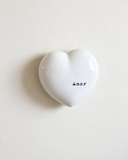 Peça decorativa Coração Declarado em cerâmica branca brilhante, com a palavra Amor escrita em preto, sobre fundo off-white.