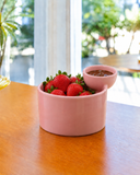 Bowl com molheira em cerâmica rosa servindo morangos, com calda de chocolate na molheira acoplada, sobre mesa de madeira e jardim ao fundo