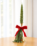 Castiçal de Natal Encanto de Natal em metal dourado com base em formato de sino, laço de tecido vermelho e pendente de ramos verdes com frutinhas, com vela em formato de pinheiro ilustrativa, sobre mesa de madeira diante de cortina clara.