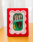 Porta retrato Sardine 13x18 cm com moldura vermelha e borda ondulada rosa, em pé sobre mesa de madeira diante de cortina clara