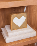 Quadro decorativo Amor nas Entrelinhas com coração em fios de linho off-white sobre fundo de linho caramelo e moldura de madeira, apoiado sobre livros em estante de madeira.