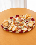 Petisqueira de cerâmica Oval Bistro Chess com borda em xadrez vermelho e off-white e dois potinhos com torradas, servida com presunto cru, queijo gorgonzola e mel sobre mesa de madeira.