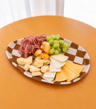 Bandeja oval de cerâmica Bistro Chess com borda em xadrez ocre e off-white, servida com salame, queijos, uvas verdes e pimenta biquinho sobre mesa de madeira.