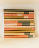 Quadro decorativo Enlace Frisos de 21x21 cm em madeira e linho, com faixas horizontais em relevo nas cores terracota, rosa claro, mostarda, areia e verde musgo, apoiado em prateleira clara.