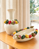 Vaso de cerâmica Tutti Frutti Oval off-white com frutas coloridas em relevo, ao lado do Vaso Tutti Frutti alto da mesma linha, sobre mesa de madeira diante de cortina de linho clara.