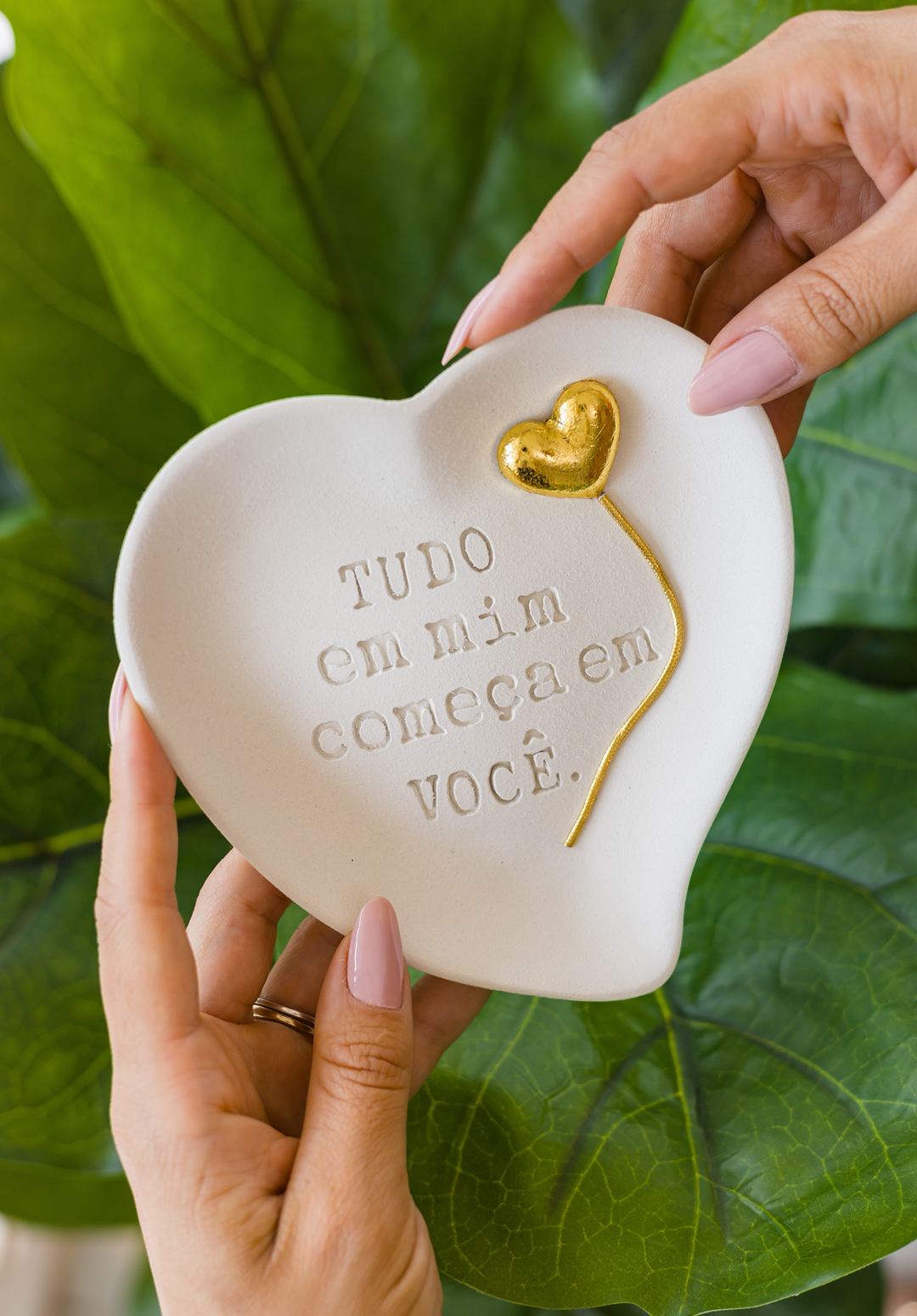 Mãos segurando peça em cerâmica fria em formato de coração com a frase tudo em mim começa em você e coração dourado, com folhagem ao fundo