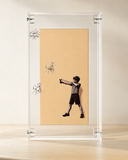 Quadro decorativo Estilingue de 26x14 cm em acrílico transparente, com foto em preto e branco de menino mirando um estilingue sobre fundo caramelo e trincas de vidro desenhadas, apoiado sobre superfície de madeira clara.
