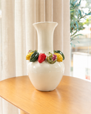 Vaso de cerâmica Tutti Frutti off-white de 42 cm com coroa de morangos, limões, figos e pimentões em relevo, sobre mesa de madeira diante de cortina de linho.