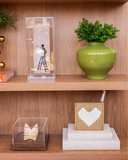 Quadro decorativo Amor nas Entrelinhas com coração em linho em estante de madeira, ao lado de cúpula de acrílico com escultura de papel, com quadro em acrílico e vaso verde com folhagem na prateleira de cima.