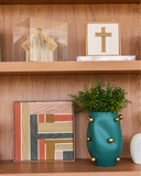 Quadro decorativo Fé com cruz em linho dourado e moldura de madeira em estante, ao lado de escultura de papel em cúpula de acrílico, com quadro de fios coloridos e vaso verde abaixo.