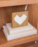 Quadro decorativo Amor nas Entrelinhas em linho caramelo e off-white com moldura de madeira, apoiado sobre livros com capa de tecido em estante.