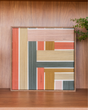 Quadro decorativo Enlace Sereno de 31x31 cm em madeira e linho, com fios em blocos de terracota, areia, verde musgo, mostarda e rosa claro, em moldura de madeira, apoiado em estante de madeira.